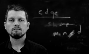 [IMAGE DESCRIPTION: a gray-scale photograph of artist, Matt Bodett is shown from the shoulders up, on the left half of the image. The right half contains the words edge, sharp, and mined in a handwritten font, white on a black background; the words are partially erased and/or scribbled out.] Courtesy of Matt Bodett.