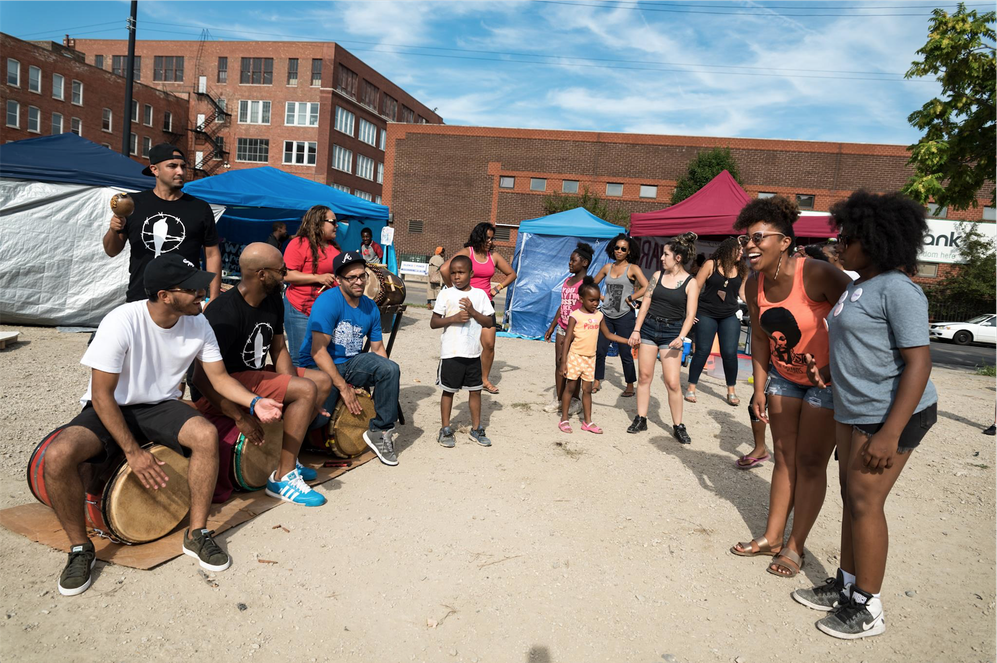 An outdoor scene in a lot with blue, white and red tents across the street from several warehouse buildings. Four percussionists at the left of the image, three sitting, two standing, playing music for an audience of people and children gathered next to them, to the right of the image.