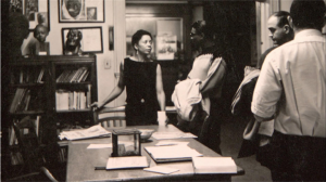 A black and white photograph of a room with framed artwork and images hanging on the walls, a bookshelf with small sculpture busts on them. Dr. Margaret T. Burroughs stands at a large table with papers on it, with three people to the left of the image talking and looking at the artwork and material in the room. Photo is takin inside of the original location of DuSable Museum on South Michigan Avenue, circa 1961. Photo courtesy of the Chicago Defender.