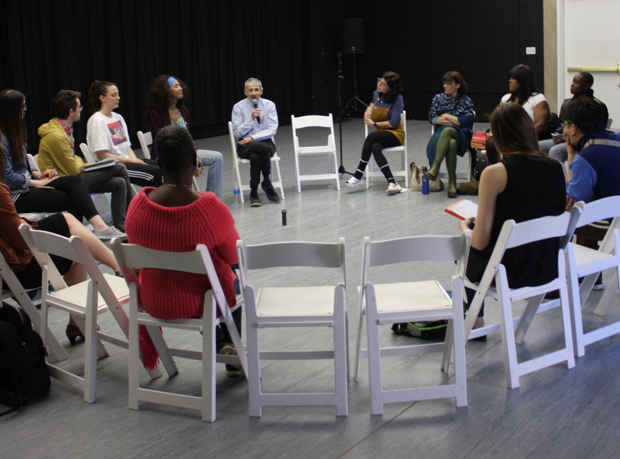 Featured image: This is a photograph of a group of people in a dance studio, sitting in a circle of chairs. Some people have their backs to the camera, and other people are shown straight-on or in profile. The two chairs nearest the camera are unoccupied, creating a window to the speaker, a man holding a microphone. Photograph by Hannah Siegfried.