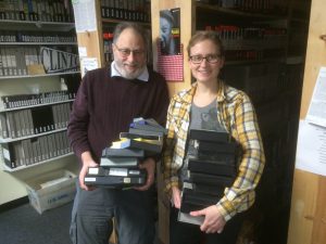 Sara Chapman and Tom Weinberg holding video tapes and moving them around as they move into their new office in 2014. They are standing in front of shelves holding more video tapes from the collection. Image courtesy of Media Burn.