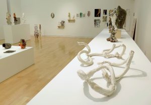 This photo shows part of the gallery. On the right-hand side of the image, a long white table stretches from the foreground toward the back wall, with the work “Lazo 018” in the foreground, “Exquisite Nomads” at the other end, and a few sculptural objects in between. “Lazo 018” is comprised of four thin, serpentine, clay objects in two pairs (one of oblong loops leaning against each other without intersecting; one of a non-looped piece reaching into and up through a closed loop). Beyond the table and along the back wall are several two-dimensional works (leaning against or hung on the wall) and three-dimensional works (on small shelves or the floor). On the left-hand side of the image, in the background, are a tall pedestal with three objects and a short platform (in partial view) with three objects visible.