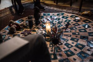 Atop a decorative mat sits a lit candle, a cup of writing instruments, some symbolic images, talking pieces, and small statues forming the centerpiece of a circle at the start of a planning meeting for teaching artists for Envisioning Justice at Circles and Ciphers.
