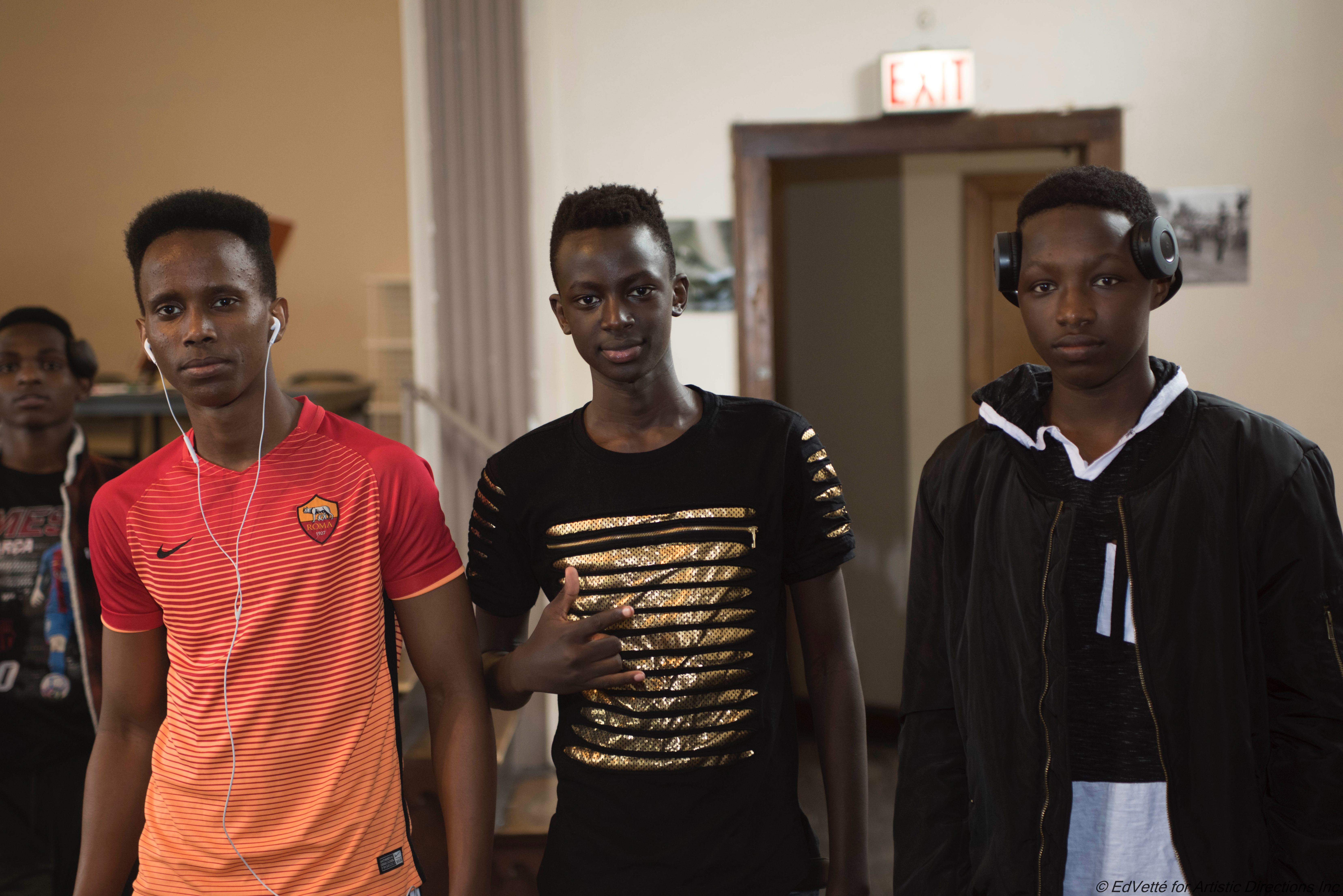 Three male students from Circles and Ciphers pose in observation of the camera: the left subject furthest to the left wears an orange and white soccer style jersey; the center subject wears a black and gold shirt while gesturing with his hand; the right subject wears a jacket and has on a large set of headphones.