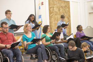 Tellin' Tales Theatre's Young Adult Writers program 2017, courtesy of Chicago Public Library. Image description: eleven people reading from black folders. Three people stand at the back while the rest are seated. Several of the participants are using wheelchairs.