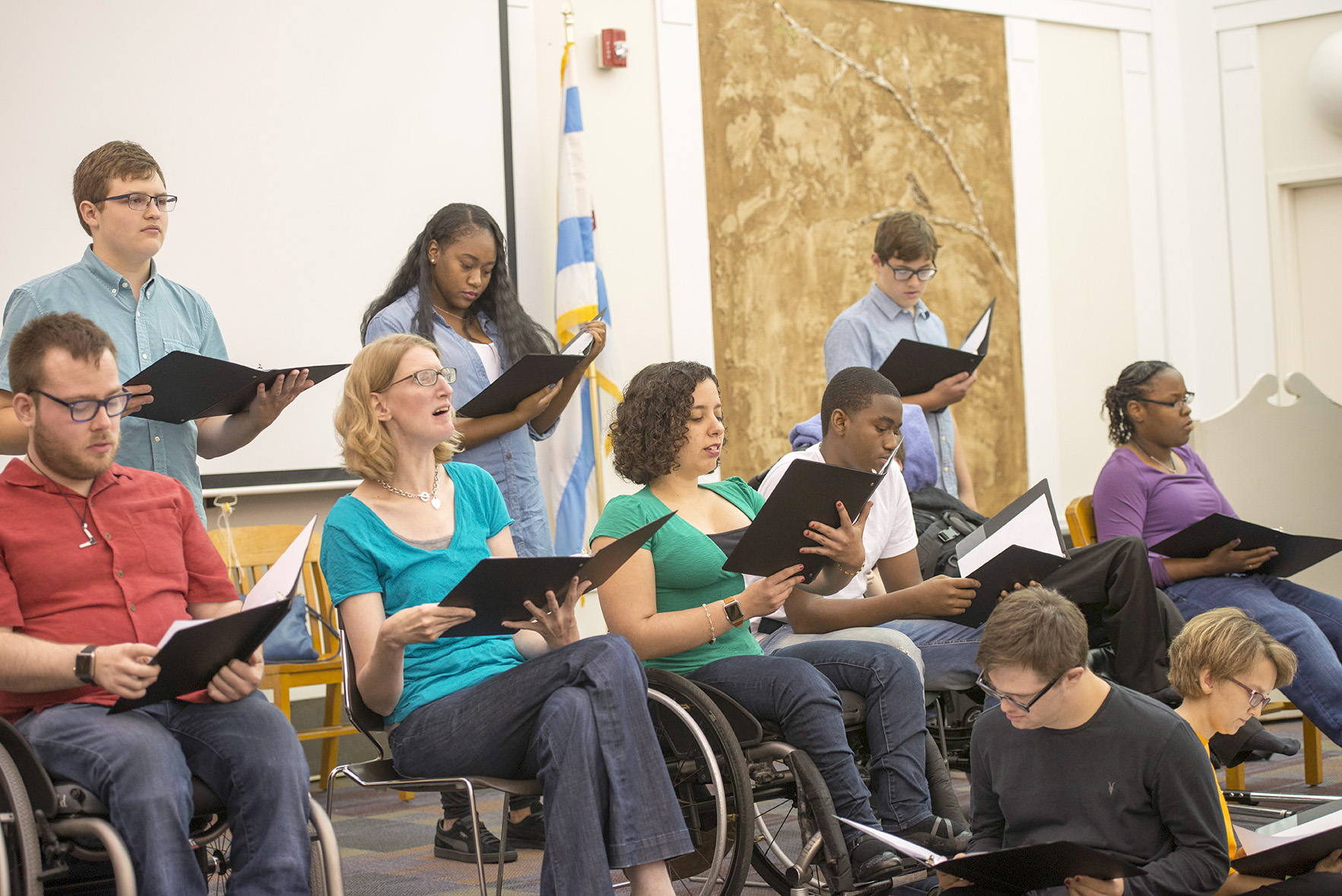 Tellin' Tales Theatre's Young Adult Writers program 2017, courtesy of Chicago Public Library. Image description: eleven people reading from black folders. Three people stand at the back while the rest are seated. Several of the participants are using wheelchairs.