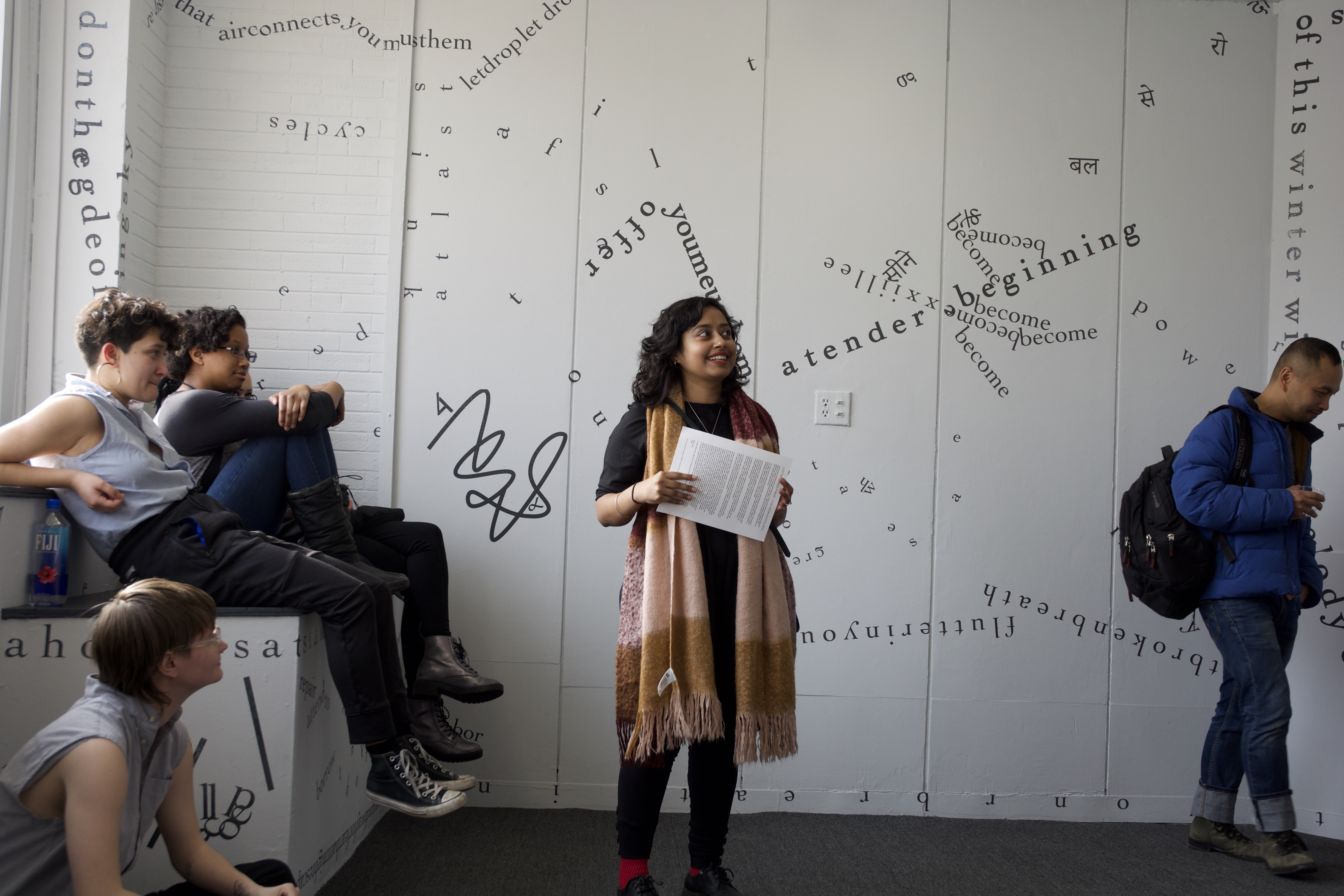 The photograph shows the artist at center, standing in front of one of the gallery’s internal, white walls, with performers and guests sitting or standing on either side of her. Black vinyl letters are installed directly onto the walls, in the form of words and phrases in English and Hindi. Text appears in different sizes and spatial orientations (e.g., right-side up, upside-down, diagonal, vertical, and organic shapes), with some words/phrases expanded in space, condensed, or intersecting with other text. English words/phrases shown in this image include “a tender beginning,” “offer,” and “of this winter.” A gestural drawing—also made of black vinyl—is shown on the left-hand side of the image.
