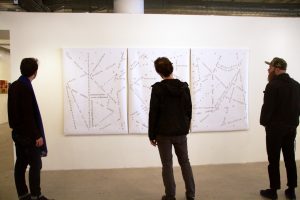 The photograph shows an interior white wall of a gallery space, on which three large pieces of canvas hang unframed and immediately adjacent to each other. Three people stand with their backs to the camera, looking at the canvases. On and across the canvases are words and phrases (written in black) that are oriented in various directions, curve, intersect, and more.