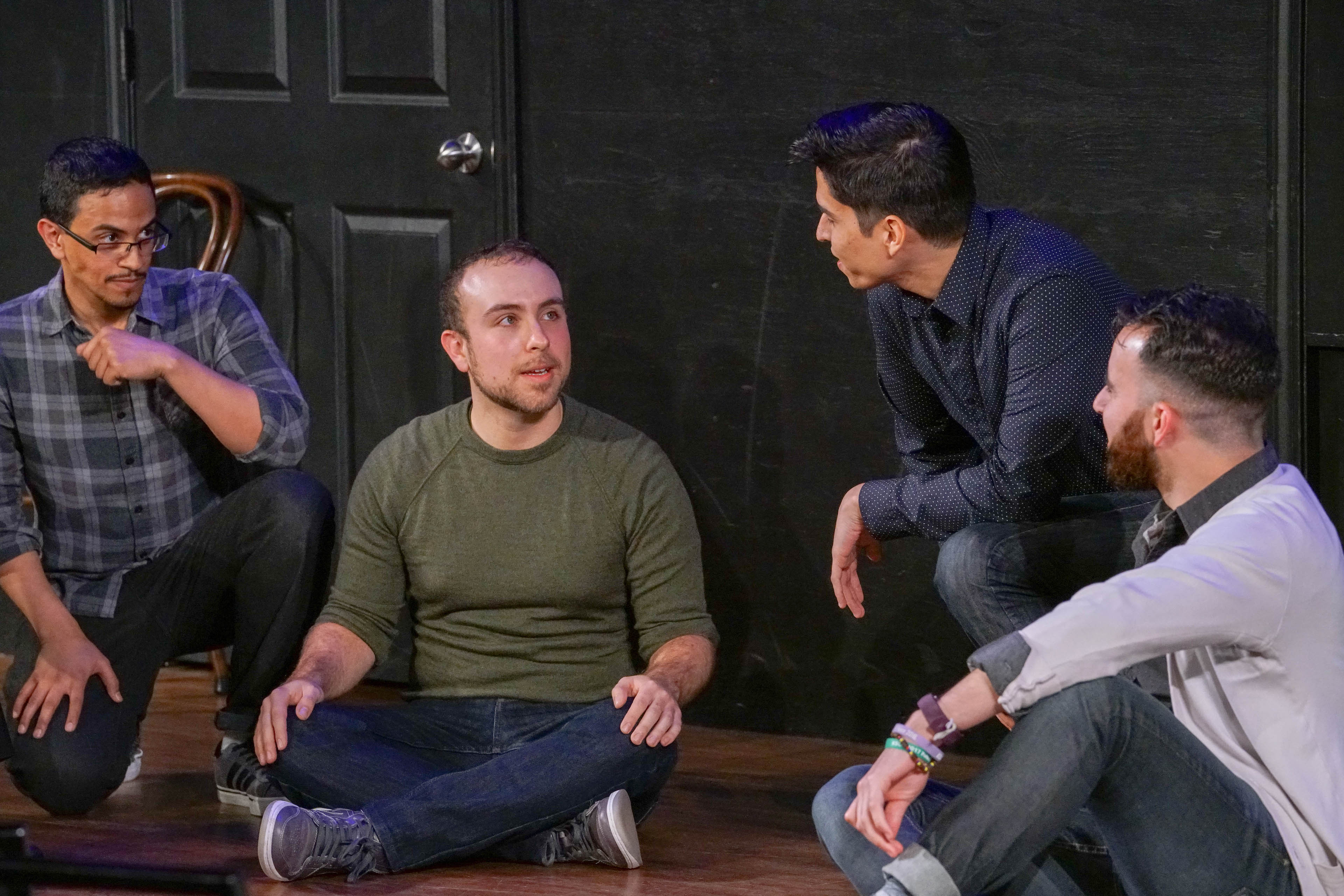 The performers sit or crouch on the stage’s wooden floor. Adam, Paul, and Devin look at Alex, whose body is angled toward the camera. Behind the performers is the black wall of the stage, with a part of a black door visible in the frame.