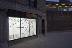 This is an external shot of the gallery, with the CTA Red Line “Loyola” elevated stop in the background. It is dim outside, and the gallery’s lights are on inside. Visible through the gallery’s large front window (which is framed by a decorative, stone façade) is the inside of the gallery—bright white walls and fixtures, with black vinyl lettering (English and Hindi) and shapes (triangular and gestural).
