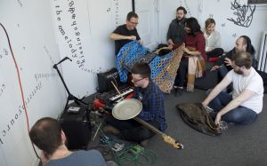 This photo is a medium-long shot, looking down toward several people sitting in a corner of the gallery. Jerry Bleem smiles while crocheting a large blue, grey, and tan piece. Udita holds the other end of it and smiles. In the foreground, a person sits holding an electric guitar, drum stick, and metal lid, with amplifiers and a keyboard nearby. Other people sit around and seem to speak to each other. Behind them are white walls and a white door onto which black vinyl letters are directly installed, in the form of words and phrases in English and Hindi. Text appears in different sizes and spatial orientations (e.g., right-side up, upside-down, diagonal, vertical, and organic shapes), with some words/phrases expanded in space, condensed, or intersecting with other text. A gestural drawing — also made of black vinyl — is shown near the center of the image.