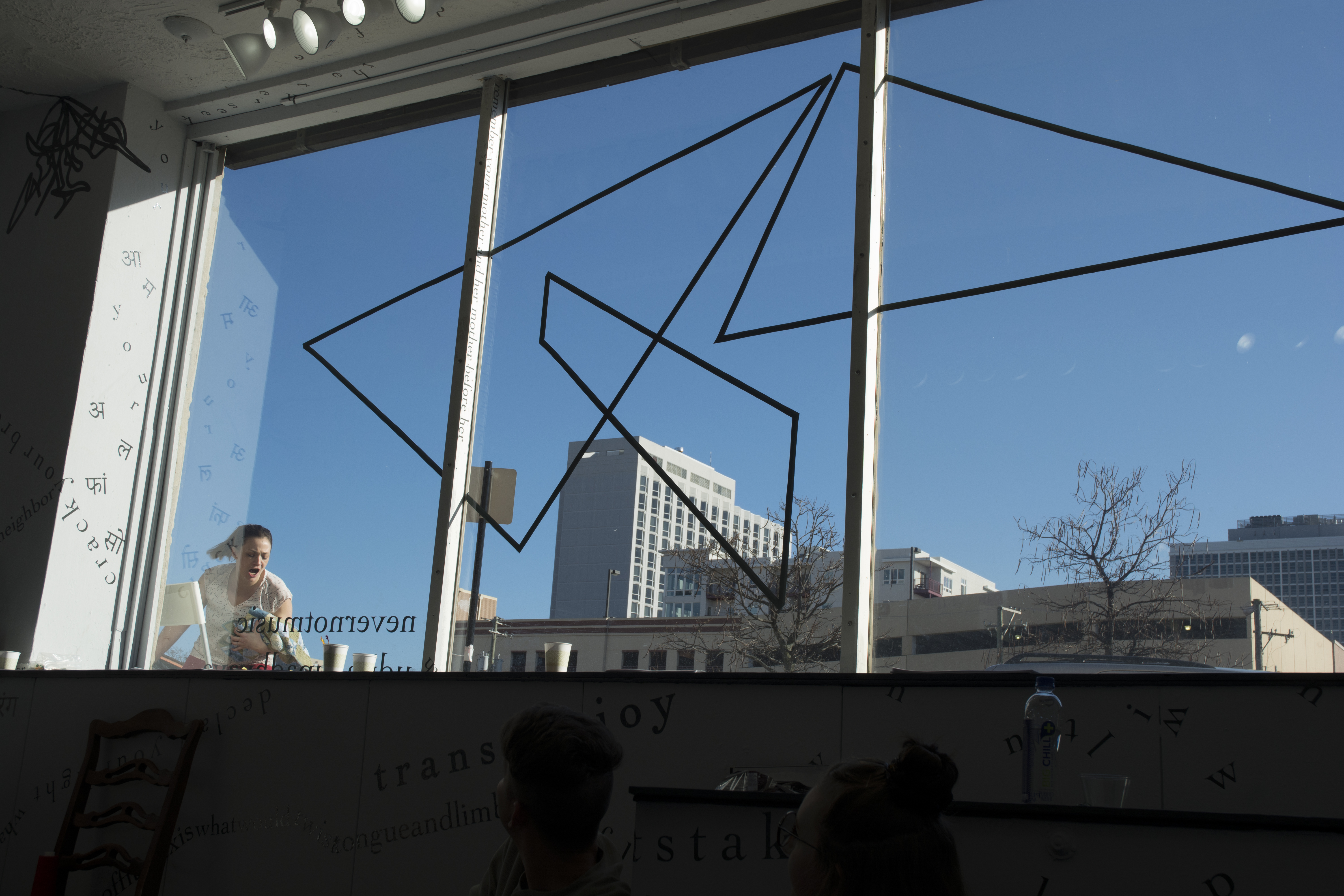 This photo shows the gallery’s large front window (with a motif of intersecting triangular shapes made of black vinyl) from the inside, and the blue sky beyond. Outside, the performer is visible through the bottom-left corner of the window. She holds something in her left arm as she moves toward the window, seemingly screaming. Inside, around the edges of the photograph, black vinyl letters are installed directly onto the white gallery walls, in the form of words and phrases in English and Hindi. Text appears in different sizes and spatial orientations (e.g., right-side up, upside-down, diagonal, vertical, and organic shapes), with some words/phrases expanded in space, condensed, or intersecting with other text. Gestural drawings—also made of black vinyl—are shown on the top left-hand side of the image. Low and very shadowy in the foreground are two audience members, sitting on the gallery floor with heads twisted toward the performer outside.