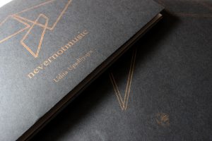 The image shows two possible versions of the book’s cover, one laid on top of the other. The version on top shows most of the cover image (thin gold parallel lines forming two triangles and a rhombus) and “nevernotmusic” and “Udita Upadhyaya” in gold text, on black paper. The book is very slightly ajar, such that the edges of its pages are visible (seemingly alternating in color, grey or white and black). The version below shows part of the same cover image, but larger and more spread out, with a tiny “Match Books” logo below it, also in gold.