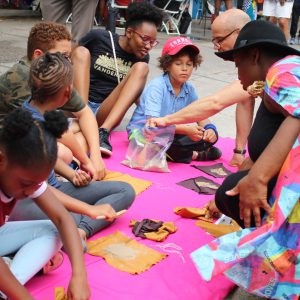 Image: Wisdom Baty (right) wears a brightly colored cape and shows a group of children how to fill nylon materials with sand at Back Alley Jazz. Image courtesy of the Museum of Vernacular Arts.