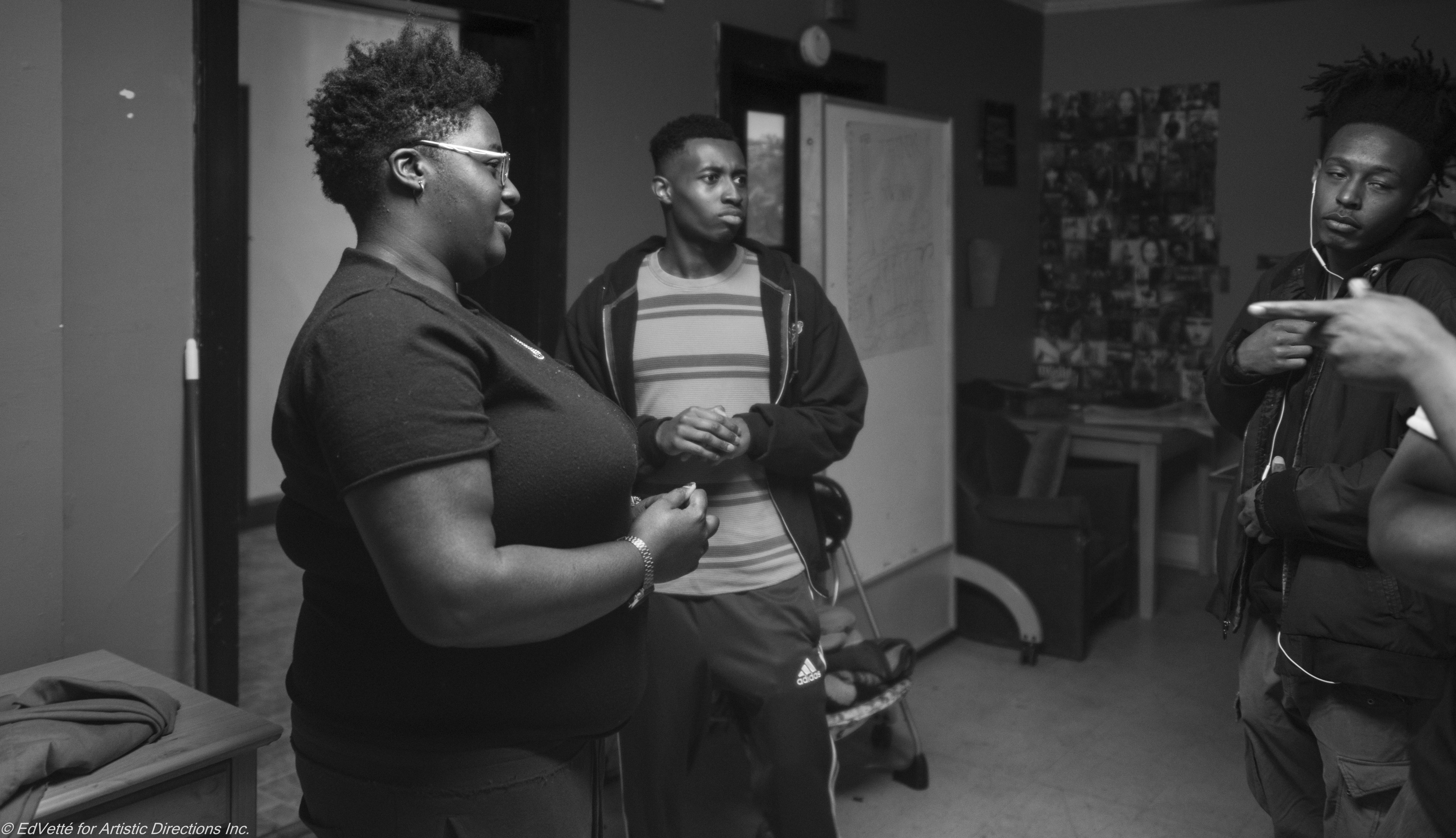 IMAGE: A black and white photo of AnnMarie Brown in conversation with three teaching artists from Circles & Ciphers. One of the artists points towards the others from out of the right frame of while AnnMarie and the two other artists look in their direction.