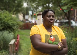 IMAGE: AnnMarie Brown stands in front of a garden area near the entrance of United Church of Rogers Park. She is staring away from the camera with arms slightly folded wearing a yellow shirt with a red rose imprint on the front.
