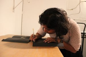 Featured image: Udita Upadhyaya at the book release for “nevernotmusic,” at TriTriangle. The artist leans over a table, looking down as she writes in gold pen inside a copy of her book. Next to her is another copy, open to its centerfold, where gold thread is visible. The artist wears a light-colored, textured sweater. Photo by Caleb Neubauer.