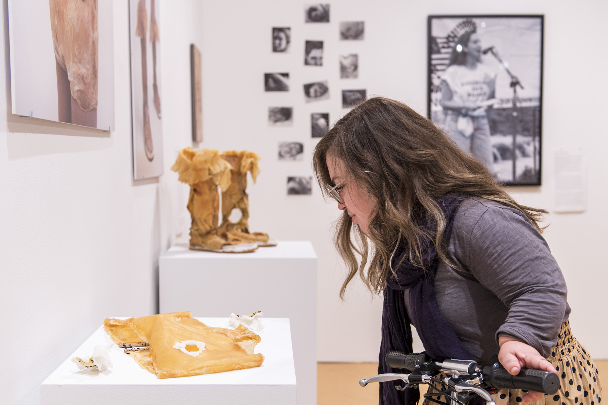 Image: Bri Beck leans into the frame from the right side, looking down at a tan mixed media garment piece on a white pedestal. Other works can be seen in the background. Photo by Ryan Edmund.