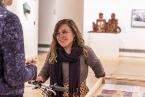 Image: Bri Beck seen in the galleries, wearing a grey sweater and dark scarf; the handlebars of her motorized scooter are visible; Courtney Graham's arm edges into the frame from the left as the two of them are in coversation. Photo by Ryan Edmund Thiel.