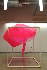 Image: a large pink semi-inflated cube sits atop a white PVC structure; it droops to the left. "Negotiating Space: Othered by Design" by Bri Beck, courtesy of the artist.