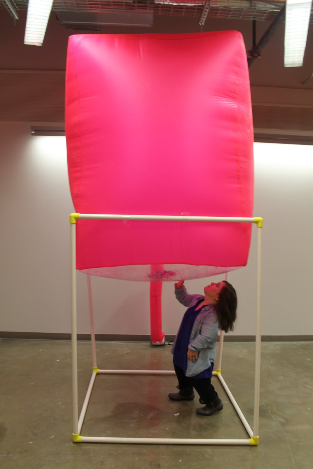 Image: a large pink inflated cube sits atop a white PVC structure; Bri Beck stands beneath it. "Negotiating Space: Othered by Design" by Bri Beck, courtesy of the artist.