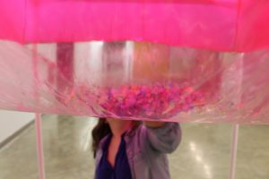 Image: a large pink inflated cube fills the top half of the frame, it is semi-transparent and confetti is seen inside; Bri Beck is on the other side of the work, touching it with her hand, her face is not visible. "Negotiating Space: Othered by Design" by Bri Beck, courtesy of the artist.