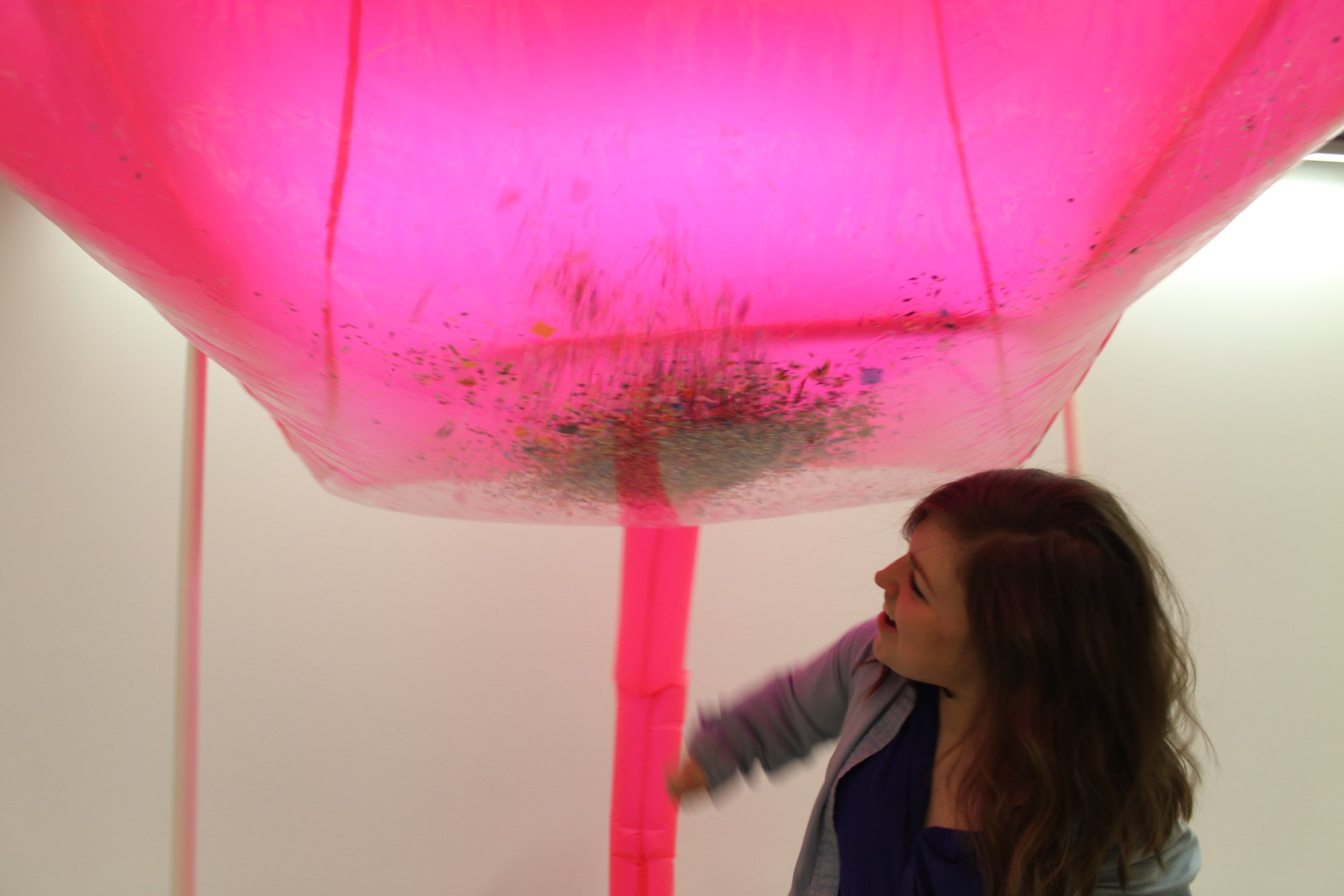 Image: a large pink inflated cube fills the top half of the frame, fed by a central tube; Bri Beck is seen beneath it in the bottom right corner. "Negotiating Space: Othered by Design" by Bri Beck, courtesy of the artist.