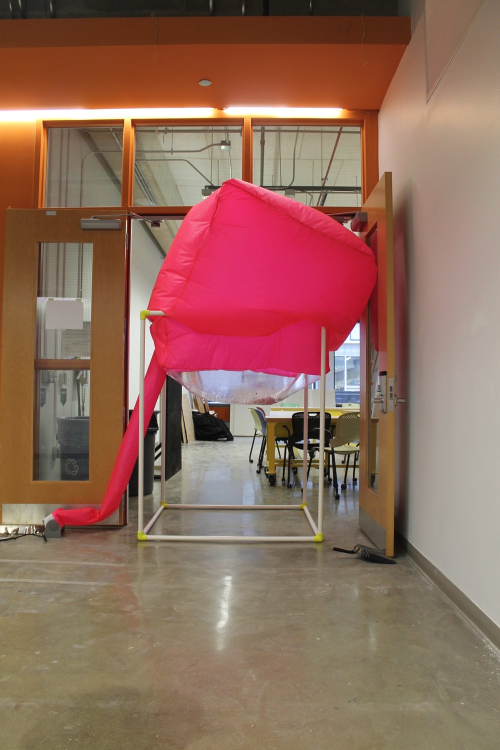 Image: a large pink inflated cube sits atop a white PVC structure; it is positioned in a doorway. "Negotiating Space: Othered by Design" by Bri Beck, courtesy of the artist.