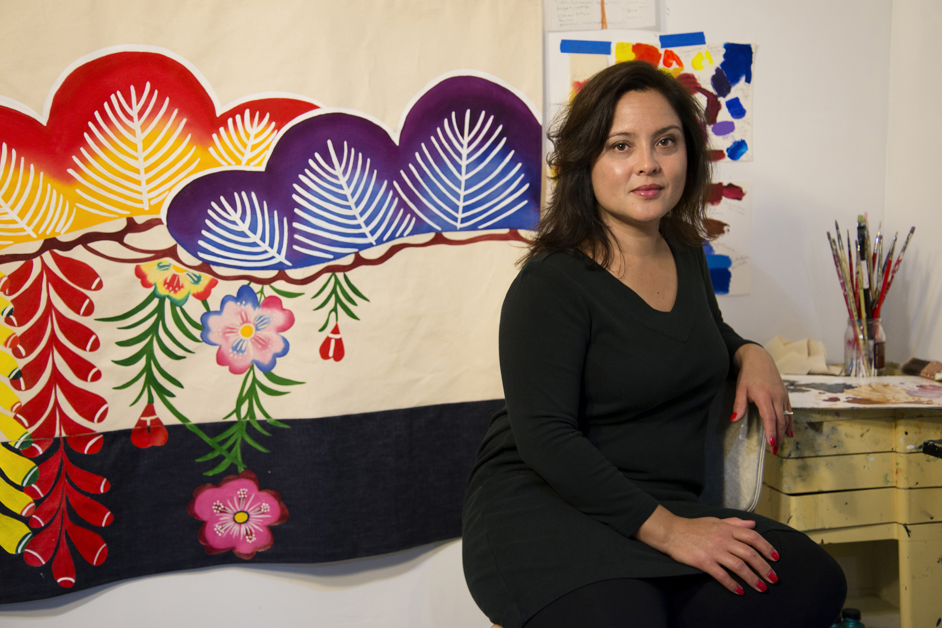 Image: Laura Kina sits in front of a brightly colored painting of floral motifs. Photo by Kiam Marcelo Junio