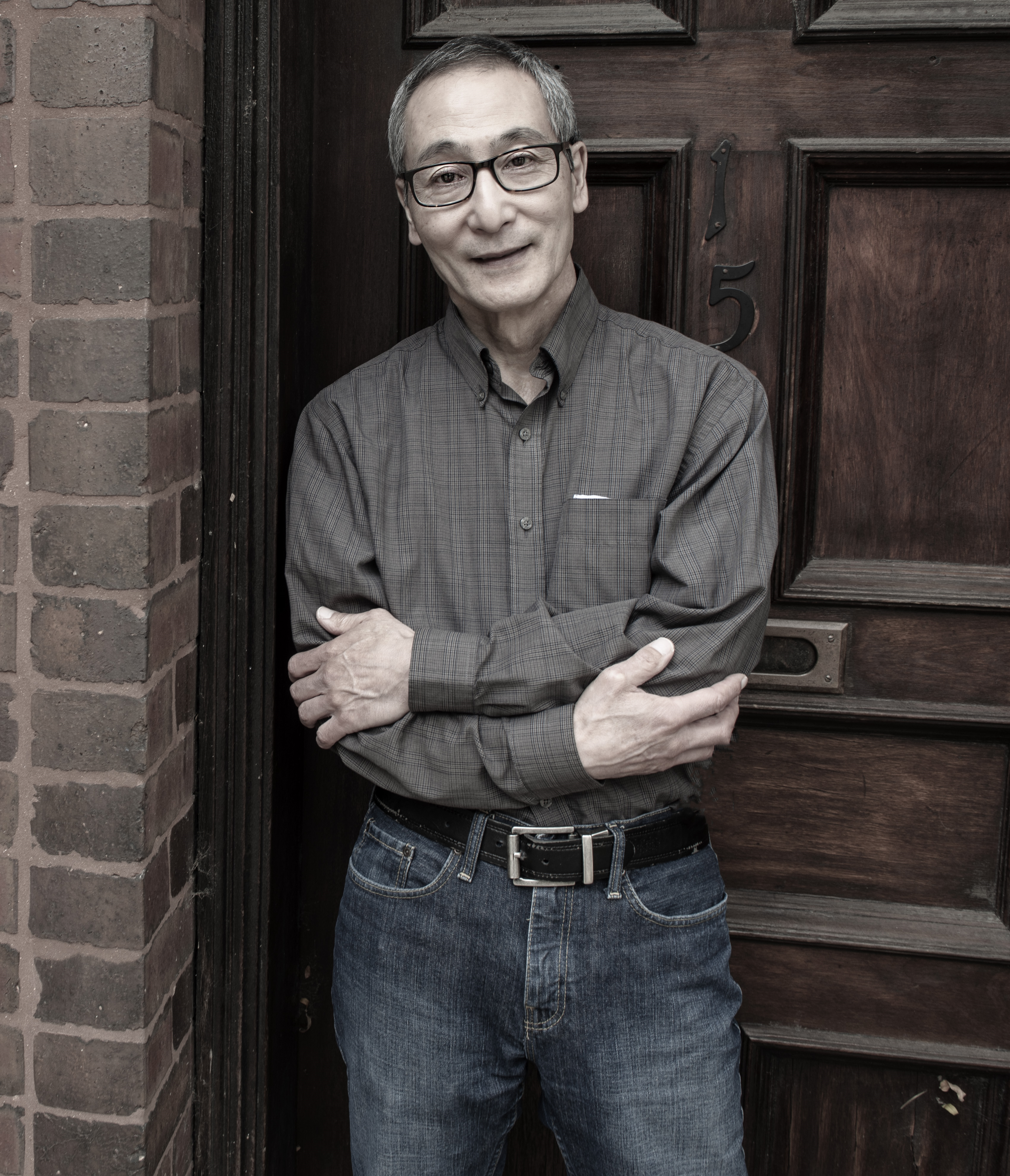 IMAGE: Steve Serikaku stands in front of a wooden door with arms folded and bearing a partial smile. He is wearing a gray button down shirt and blue jeans. Photo by Edvetté Wilson Jones.