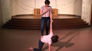 Featured image: Maggie Robinson and Allison Sokolowski performing in “I Am” at the Chicago Danztheatre Auditorium, as part of the Body Passages culminating event. Maggie balances with one foot, knee, and hand on the floor, as Allison stands on Maggie’s lower back. The performers hold each other’s left hands and look at each other. Both are barefoot and wear white t-shirts and jeans. Behind them is a well-lit stage, with a string of colorful paper suspended across it. Still from a video by John Borowski.