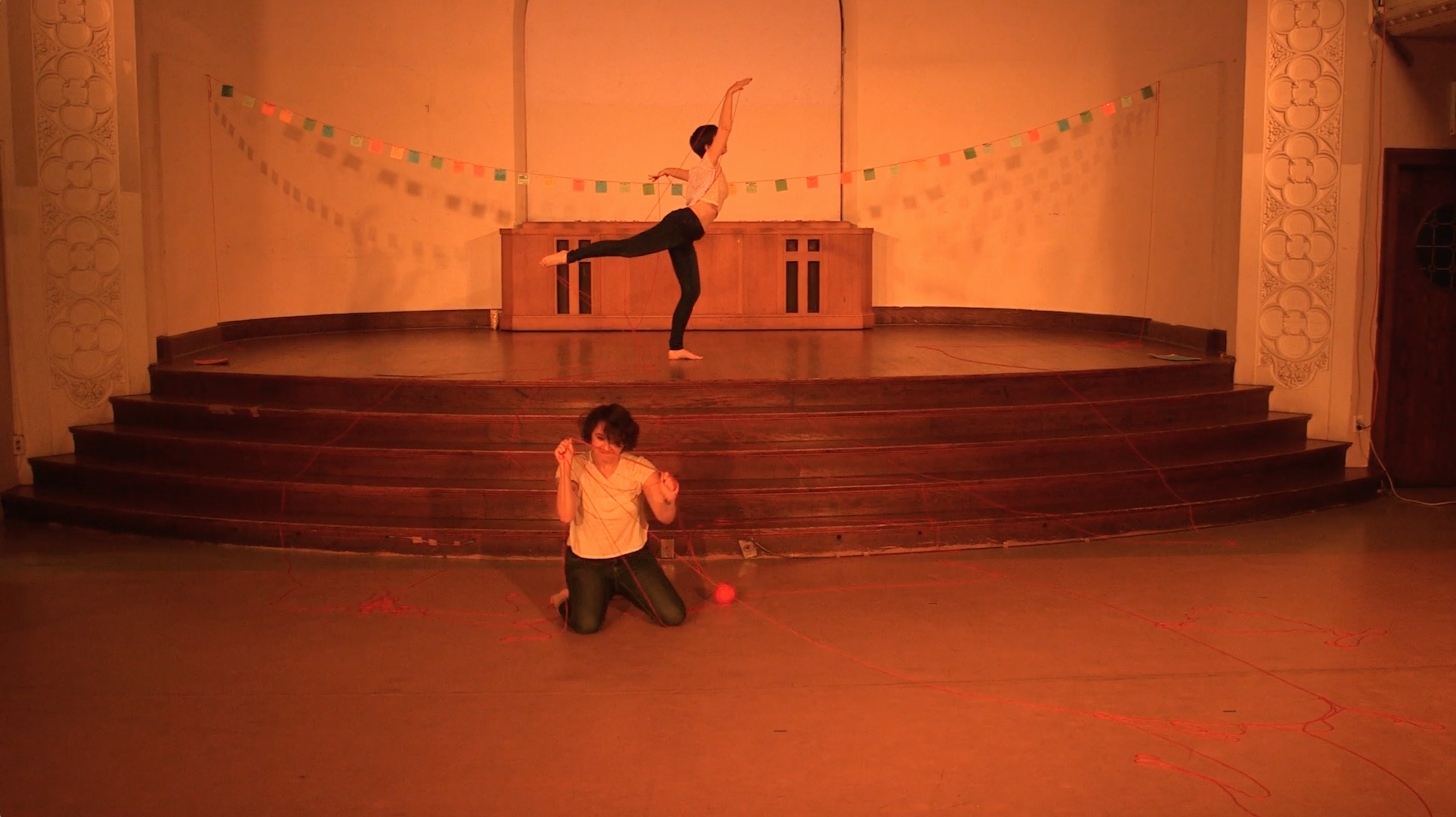 Image: Maggie Robinson and Allison Sokolowski performing in “I Am.” In the foreground, Maggie kneels on the floor, holding up strands of red yarn, which is also strewn across the floor. In the background, Allison stands onstage in an arabesque, also holding up red yarn. Both performers are barefoot and wear white t-shirts and jeans. The stage is bathed in warm orange light and has a string of colorful paper suspended across it. Still from a video by John Borowski.