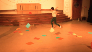 Image: Allison Sokolowski performing in “I Am.” In this long shot of the performance area, Allison runs with arms spread as colorful pieces of paper fall through the air and onto the floor. A shadow of Maggie in a similar pose (off-camera) is slightly visible. The performance space is bathed in warm yellow-orange light. Still from a video by John Borowski.