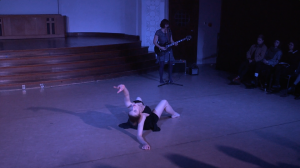 Image: Carly Broutman and Michelle Shafer performing in “Wax.” In the back corner, Michelle plays electric guitar and sings into a microphone on a stand. Carly is on the floor, body and feet angled toward Michelle, while leaning back on one elbow, reaching and looking past the camera. The audience is partially visible in the background and via cast shadows in the foreground. The lighting is dim and cool (blue-purple). Still from a video by John Borowski.
