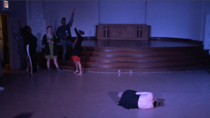 Image: Carly Broutman and Jae Green performing in “Wax.” Carly lies on the floor in a ball in the foreground, her back to the camera. In a far corner, Jae and three guests celebrate, smiling, yelling, holding beverages, and/or throwing arms in the air. The lighting is dim and cool (blue-purple). Still from a video by John Borowski.