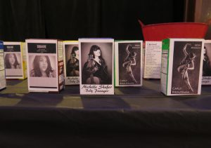 Image: The interactive installation related to Carly Broutman, Jeanette “Jae” Green, and Michelle Shafer’s performance “Wax.” This is a close-up image of a table with a black tablecloth, on which several single-serving cereal boxes sit, each stickered on one side with a black-and-white photograph of a group member. Visible in the background are the back of one box, including lyrics for “Cocooned,” and a red bucket. Photo by Marya Spont-Lemus.