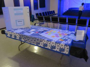 Image: The interactive display related to Lani T. Montreal and Maxine Patronik’s performance “Blood Memory,” at the Chicago Danztheatre Auditorium as part of the Body Passages culminating event. In the foreground, a table is covered with blue-and-white elephant-print cloth, a tri-fold posterboard, and various papers, including a large one that reads “SUPPORT BELIEVE