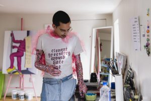 Image: The artist is in their studio wearing pink and purple gloves. They are wearing a white shirt with text that says "Clement Greenberg would hate me" and looking down at a table. In the background there is a painting on an easel with a pink figure wearing crocs. Photo by Ryan Emdund Thiel.