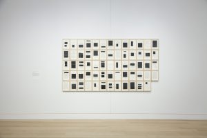 Image: Bethany Collins, Southern Review 1985 (Special Edition), 2014-15 [installation view]. 64 sheets fill a 13x5 panel with the lower-rightmost square removed. Each sheet is filled with blacked-out squares covering whatever text might appear on the page. Image courtesy of DePaul Art Museum.