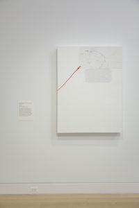 Image: Brenna Youngblood, Untitled, 2012 [installation view]. Several everyday objects, including a flattened 30-pack of beer, are painted white and plastered onto a white canvas. A red soda can tab linked to a long red line stands out. Image courtesy of DePaul Art Museum.