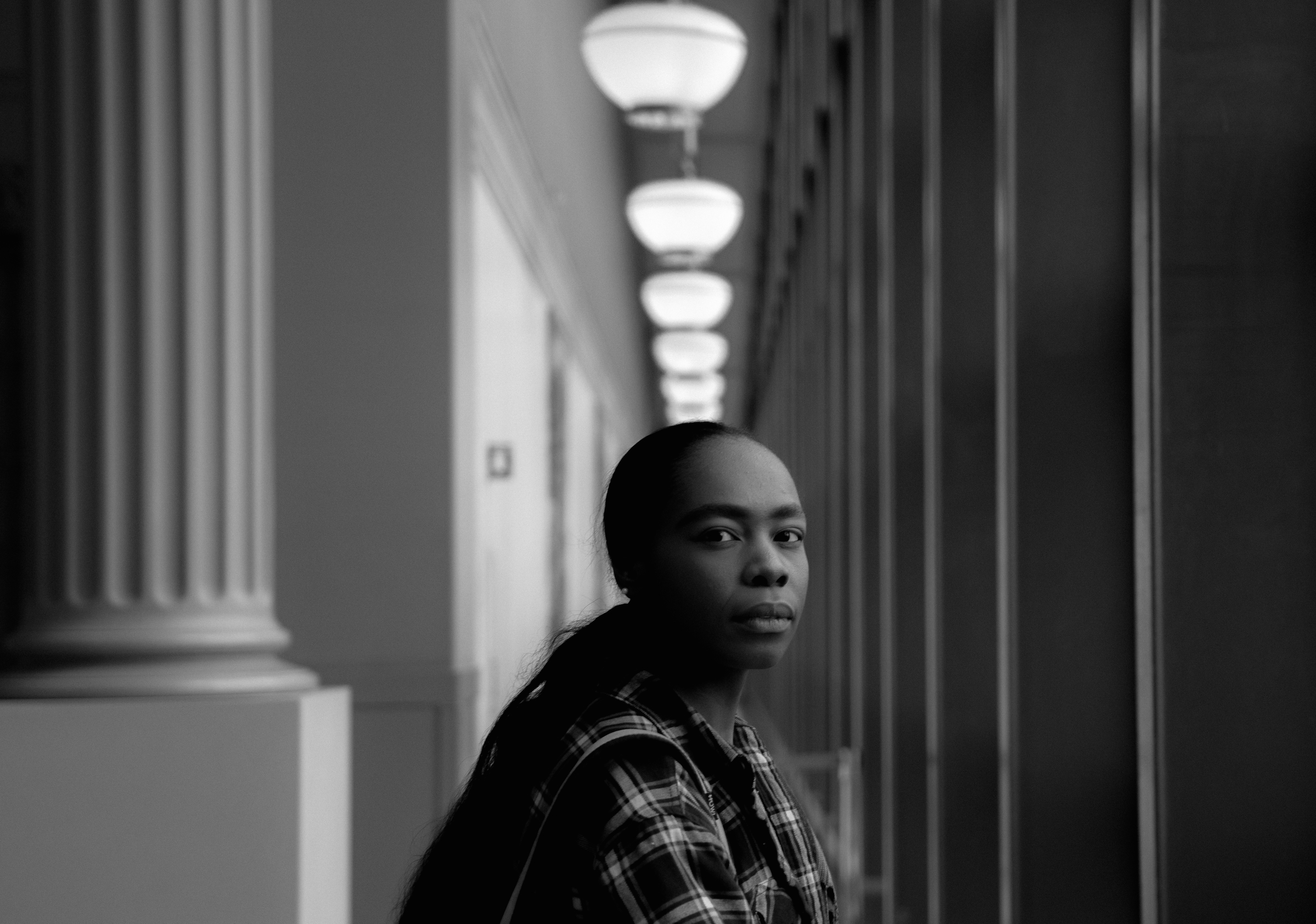 Image: Black-and-white portrait of bria royal in semi-profile, flanked on either side by columns and beams. Above her head are a series of globular light fixtures. Photo by Sebastián Hidalgo.