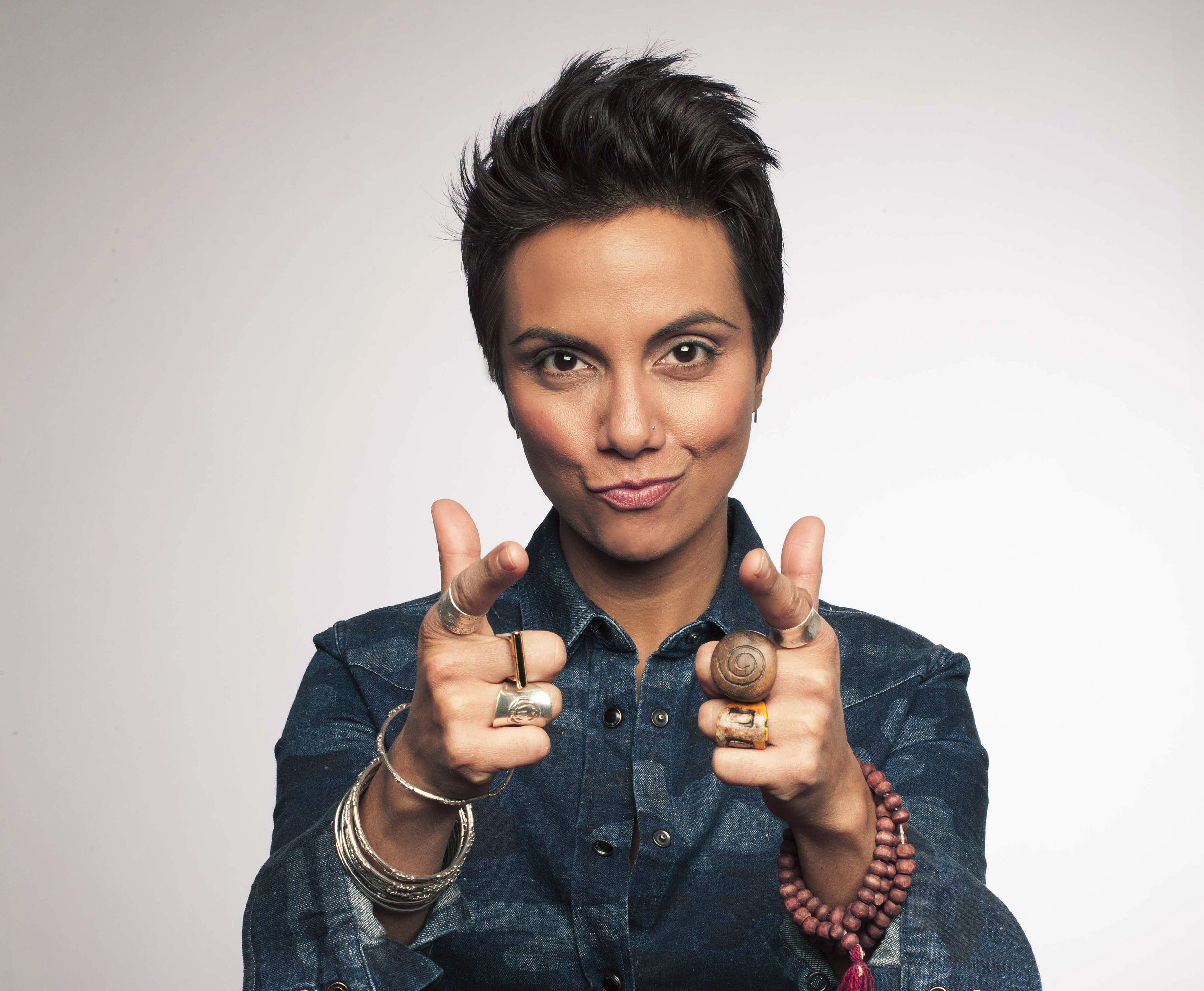 Featured image: Fawzia Mirza. In this medium-close-up shot, Fawzia looks directly and confidently at the camera, smiling with pursed lips and pointing at the viewer with both hands. Fawzia wears a long-sleeved denim shirt with a blue-on-blue, camouflage-like print and the top few snaps unsnapped. The cuffs of her shirt-sleeves are folded back, and she wears several bracelets and rings made of silver or wood. Photo by Bradley Murray, cropped to fit. Courtesy of the artist.