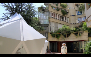 Image: Video still from Hương Ngô's, In the Shadow of the Future, 2014-19. Still shows someone dressed up in a cosmonaut outfit standing in the foreground next to a large white geometric structure. In the background is a building with plants dangling out the windows, with architecture in a similar triangular style as the white structure as well as Hương's central installation. It is a sunny day out in the photo. Image courtesy of the artist.
