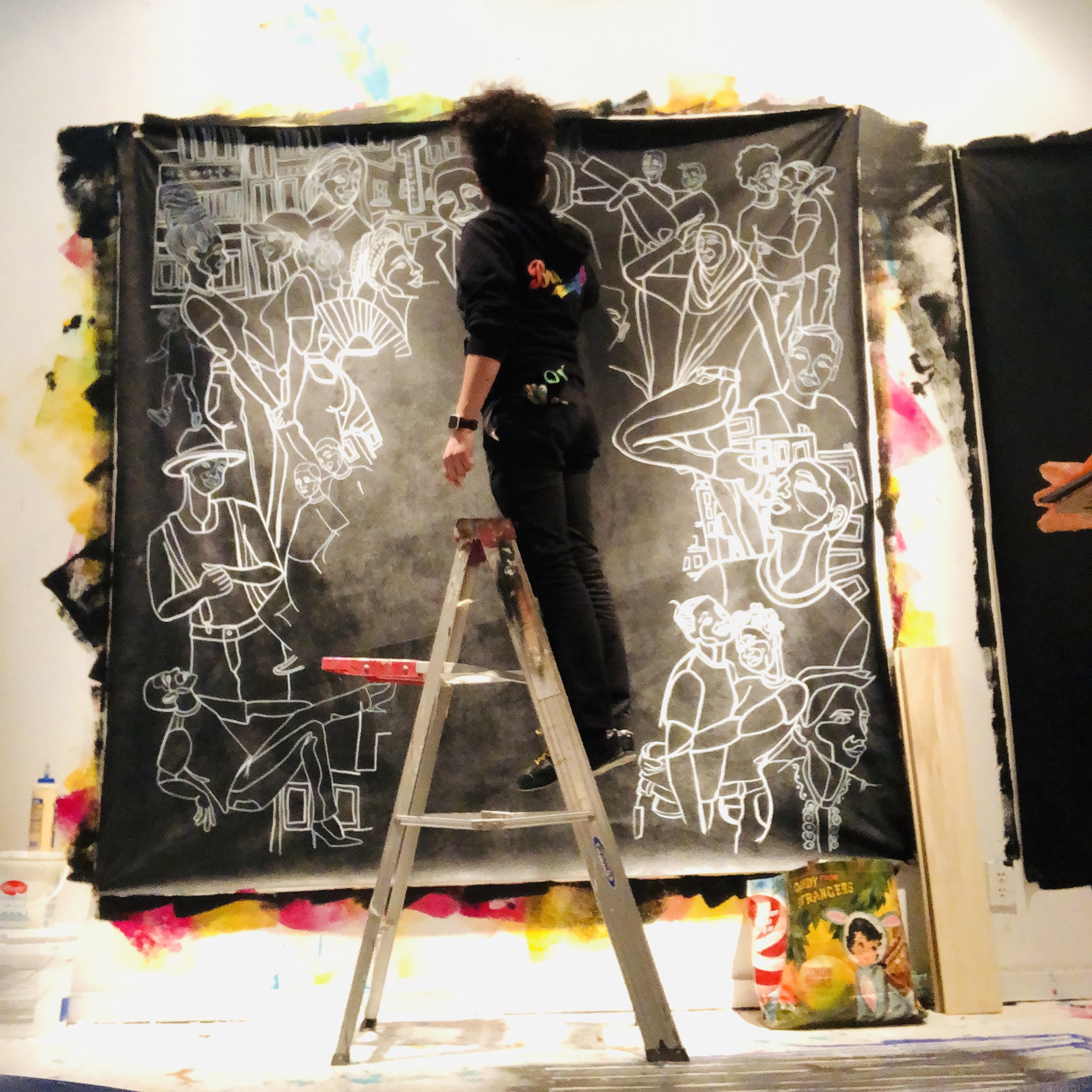 Image: The artist Sam Kirk standing on a ladder and painting a canvas in progress – a black background with white figures. Image courtesy of artist.