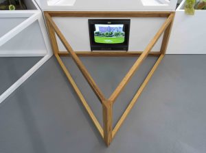 Image: Closeup photo looking down on a wooden triangular segment placed on a grey gallery floor. One of the triangle’s points faces directly at the viewer and the side farthest away is pressed against a white wall. Built into this wall, perpendicular to the floor and only a couple of inches above the ground, is a small television. The screen shows a big building behind a grassy field and a line of trees. Image courtesy of the artist.