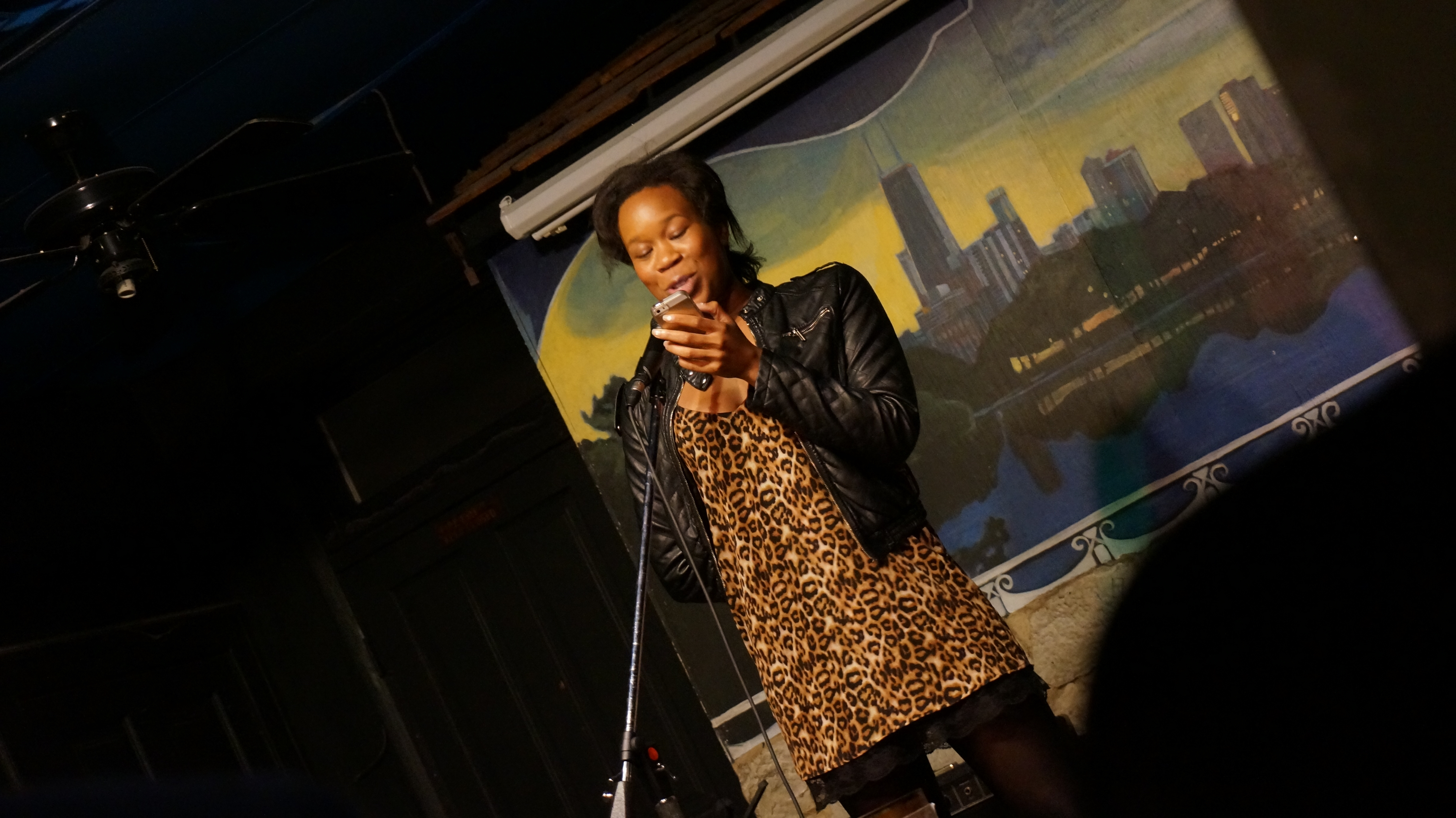 Image: Photo of Britt Julious performing on-stage at Miss Spoken at the Gallery Cabaret. Julious stands on-stage, speaking into the microphone while looking at a phone. Julious wears a black jacket over an animal-print dress with black trim. The reader appears in medium-long-shot at the center side of the frame, which is skewed at a diagonal. Painted on the stage wall behind the reader is a scene showing Chicago’s skyline, as if viewed from behind a stone wall. Photo by Sarah Joyce. Courtesy of Miss Spoken.