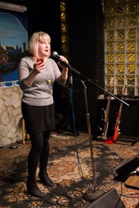 Image: Photo of Rosamund Lannin performing on-stage at Miss Spoken at the Gallery Cabaret. Lannin looks out toward the audience (not visible) and speaks into the microphone, while holding a phone in one hand. Lannin wears a grey shirt, black skirt, black leggings, and black boots, and stands on a multi-colored rug. Painted on the stage wall behind the reader is a scene showing Chicago’s skyline, as if viewed from behind a stone wall. Also visible in the background are two electric guitars, set to lean toward a black wall with glass-brick windows. Photo by [Photographer]. Courtesy of Miss Spoken.