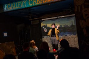 Image: Photo of Brittany Meyer performing at Miss Spoken at the Gallery Cabaret. The reader stands on-stage, smiling and speaking into the microphone while looking out at the audience. Meyer’s hands are raised in expression; one hand holds loose papers and the other is open. Meyer wears a mustard-gold cardigan open over a black turtleneck, a dark patterned skirt over dark leggings, and glasses. The reader appears small near the center of the frame. Painted on the stage wall behind the reader is a scene showing Chicago’s skyline, as if viewed from behind a stone wall. In the foreground, the words “Gallery Cabaret” are partially visible at the top of the frame and the backs of some audience members’ heads are visible at the bottom. Photo by Sarah Joyce. Courtesy of Miss Spoken.