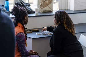 Image: Jamila Kinney (left), who led a guided sitting and walking meditation during the day's events, and event co-curator Courtney Cintrón (right) sitting a table together with their backs to the camera. Courtney is facilitating a tarot card reading for Jamila and is pointing to three stacks of cards. Photo by Ryan Edmund.