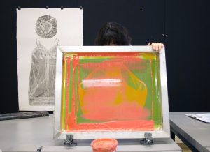 Image: Guen Montgomery holds a screenprinting screen up, exposing the neon pink paint-covered screen to the camera. The top of her head is visible above the screen. Pinned behind her on a black corkboard is a gray collagraph print of a woman’s shower cap and nightgown on white paper. Photo by Jessica Hammie.